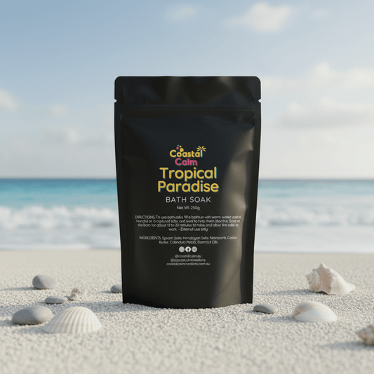 Black packaging of 'Coastal Calm Tropical Paradise Bath Soak' on a beach with shells and ocean in the background.