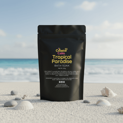 Black packaging of 'Coastal Calm Tropical Paradise Bath Soak' on a beach with shells and ocean in the background.